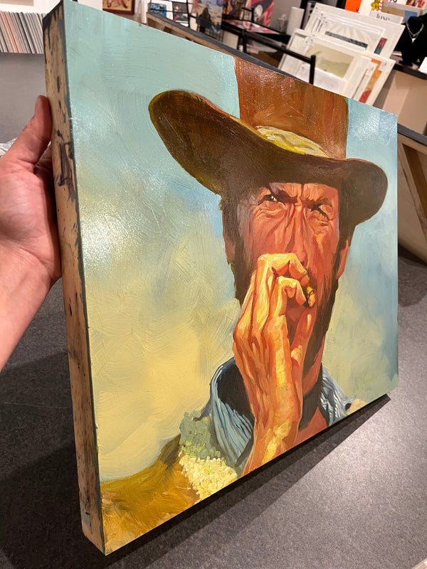 Gabe Leonard's painting of the actor Clint Eastwood wearing a cowboy hat smoking a cigar with his right hand. He is wearing a blue collared shirr and a brown cowboy hat.